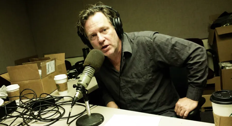 A man at a podcast microphone, shot on a phone under strip lighting, cardboard boxes and tangled cables filling the room behind him.